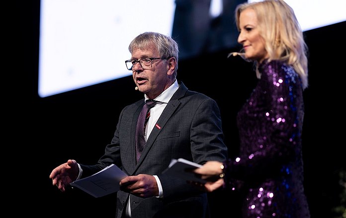 Zwei Personen stehen auf einer Bühne und moderieren eine Veranstaltung. Ein Mann im Anzug hält Unterlagen in der Hand und spricht in ein Headset-Mikrofon, während eine Frau im glitzernden violetten Kleid daneben steht.