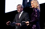 Zwei Personen stehen auf einer Bühne und moderieren eine Veranstaltung. Ein Mann im Anzug hält Unterlagen in der Hand und spricht in ein Headset-Mikrofon, während eine Frau im glitzernden violetten Kleid daneben steht.