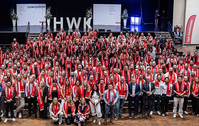 Große Gruppe von Menschen bei einer Meisterfeier der Handwerkskammer Köln im RHEIN SIEG FORUM. Viele Teilnehmende tragen rote Schals mit der Aufschrift „Handwerksmeister“ und stehen gemeinsam für ein Gruppenfoto vor der Bühne.